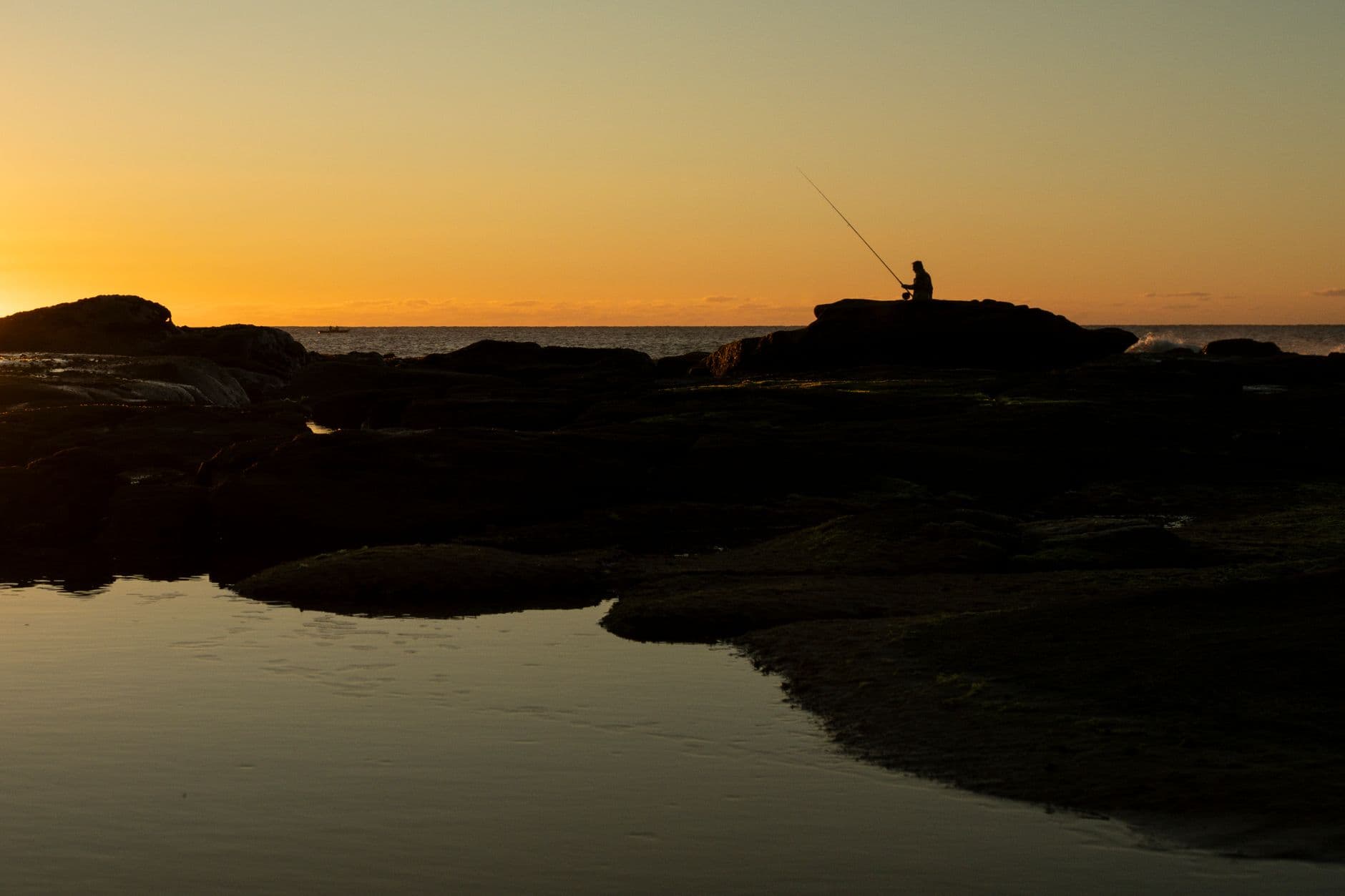 An angler casting from rocks, silhouetted against an orange sunrise over the ocean.